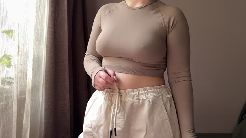 A young woman proudly shows her weight loss progress at home, holding a shaker with a healthy drink. Bright apartment setting, fitness and wellness lifestyle.
