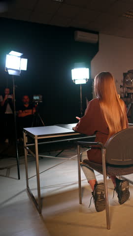 Unrecognized red-haired lady sits in the middle of the studio surrounded by soffits. Man and woman taking video working backstage. Vertical video.