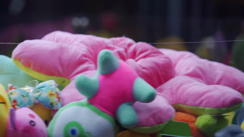 A variety of brightly colored plush toys are displayed inside a claw machine. The toys sit behind a clear glass barrier in an indoor arcade environment, ready to be won by players.