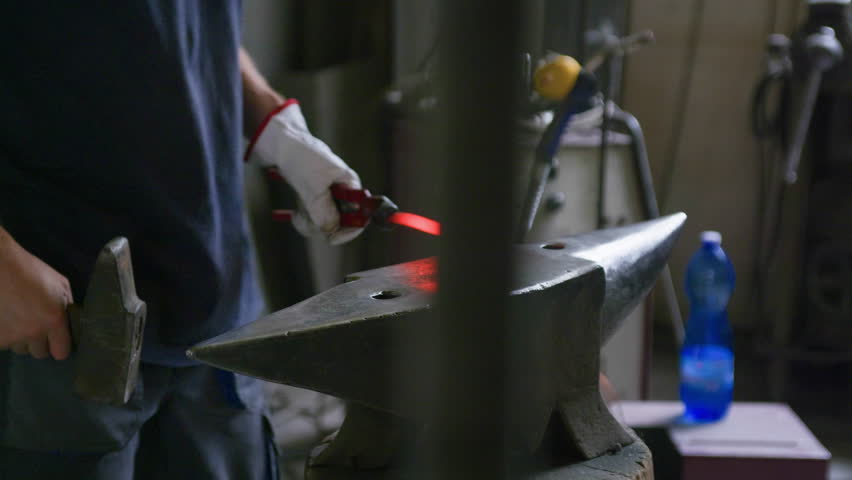 Side view of blacksmith hammering hot metal on anvil with forge bokeh in foreground. Sparks and warm lighting highlight traditional forging craftsmanship in workshop.