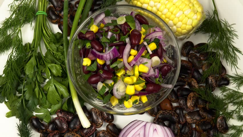 Glass bowl with vegetable salad based on corn, top view. Fresh corn on the cob, red bean, red onion, dill, parsley and green scallions