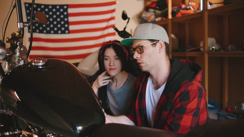 Lovely couple focus intently while repairing a motorcycle in a cozy garage adorned with an American flag, showcasing teamwork and passion for bikes.