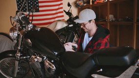A young man wearing a cap and sunglasses works on a motorcycle in a garage, surrounded by tools, another bike, and an American flag wall decoration. - Powered by Shutterstock - Get 15% off with code: PIKWIZARD15
