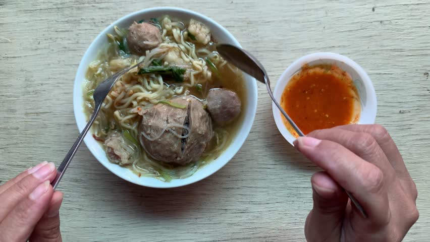 4K top view of a person eating bakso noodle soup with chili sauce on rustic table. Warm mood, traditional vibe. Perfect for food, lifestyle, culinary, street food, or Southeast Asian culture themes.