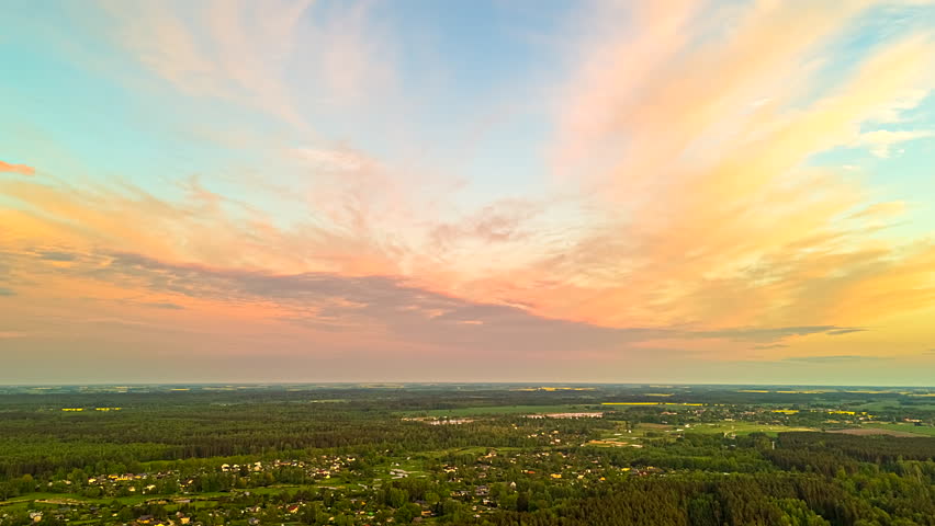 A scenic time-lapse captures a serene sunset painting the vast sky with soft pastel pink and orange clouds above a sprawling rural landscape of green forests and small villages in Latvia.
