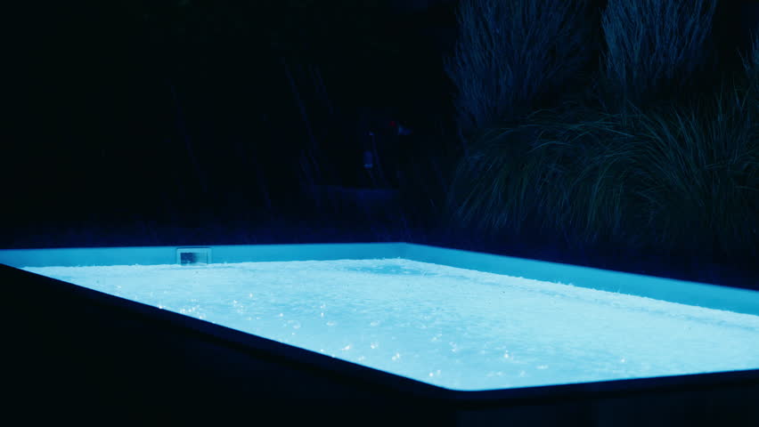 Illuminated pool at night in heavy rain with visible raindrops hitting the glowing water surface and dark foliage in the background. High quality 4k footage