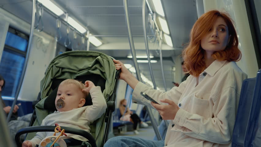 In a bustling subway, a mother holds her smartphone while tending to her child in a stroller. They travel together, showcasing daily life in the city.