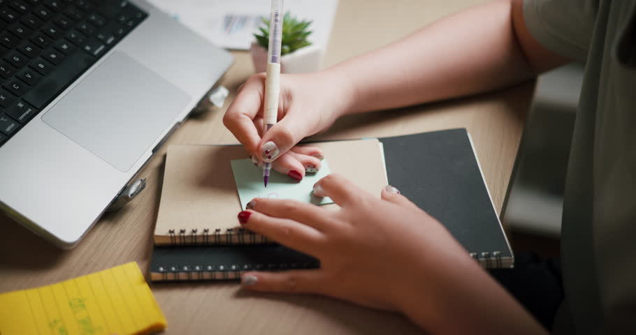 Handheld selective focus shot, Woman's hand writing message on light blue sticky note with purple pen on desk with laptop and notebook.