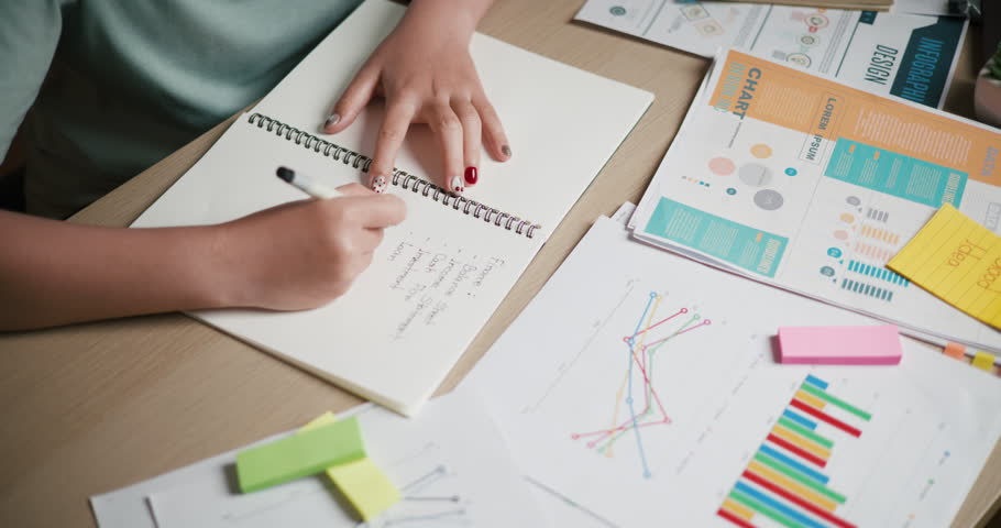 Handheld top view shot, A woman is taking notes in a notebook while sitting at a desk filled with business charts and sticky notes.