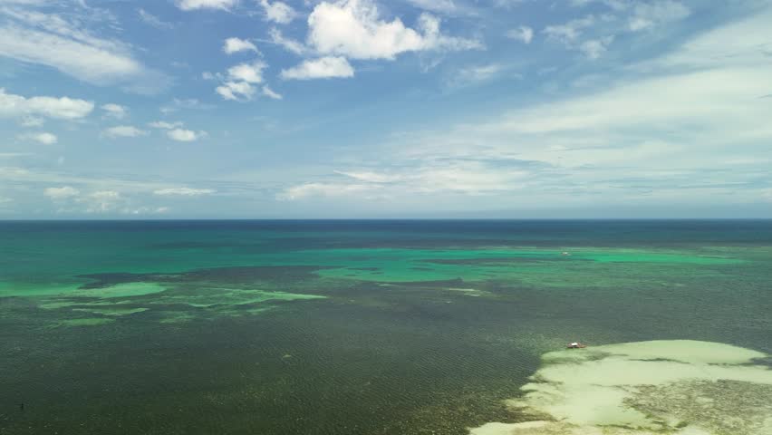 High aerial glide reveals turquoise reef flats, pale sandbars and deep blue channels off Barimbing Beach, San Jose, Puerto Princesa, Palawan, as the calm Sulu Sea meets a cloud-flecked sky.