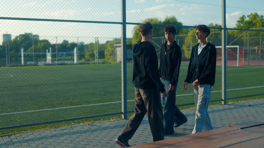 Three friends enjoy a stroll near a green sports field, deeply engaged in conversation. Hoodies on, sunlight out just another solid afternoon hangout.