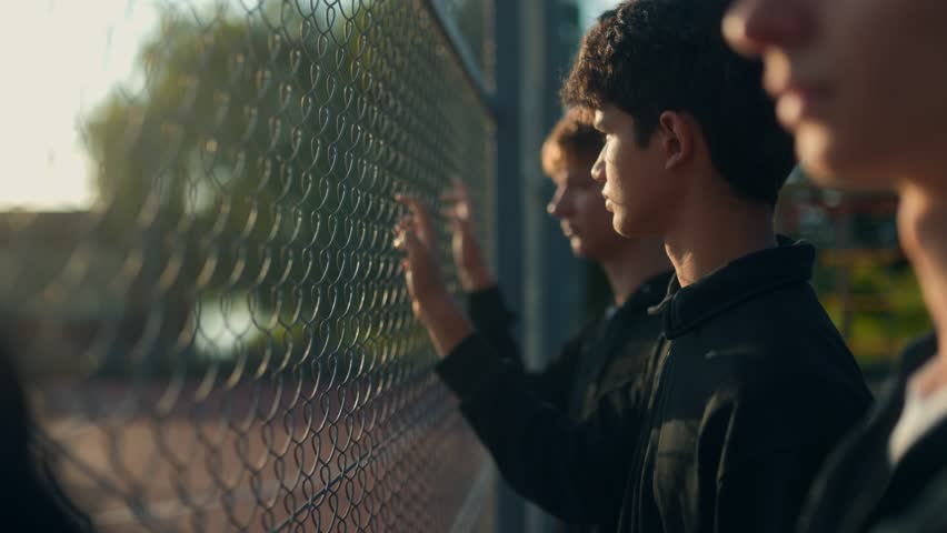 A trio of teen boys hangs by a chain-link fence under evening light, capturing a moody but heartfelt scene of close friendship, quiet reflection, and connection.