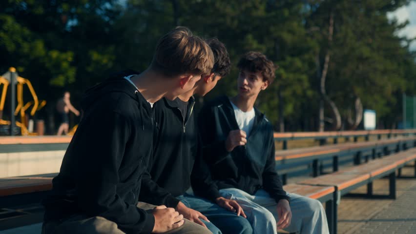Three teenage boys sit on school bleachers, chatting and laughing together under the sun. A casual scene of real friendship and fun outdoor conversation.