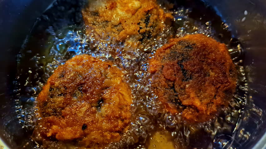An extreme close-up shot shows three homemade meat patties, similar to Latvian kotletes, deep-frying to a golden-brown crisp in a pot of hot, bubbling, and sizzling cooking oil.