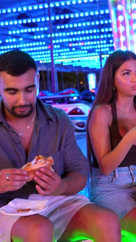 Friends sharing hot dogs under bright carnival lights, creating joyful memories during nighttime entertainment at lively amusement park destination