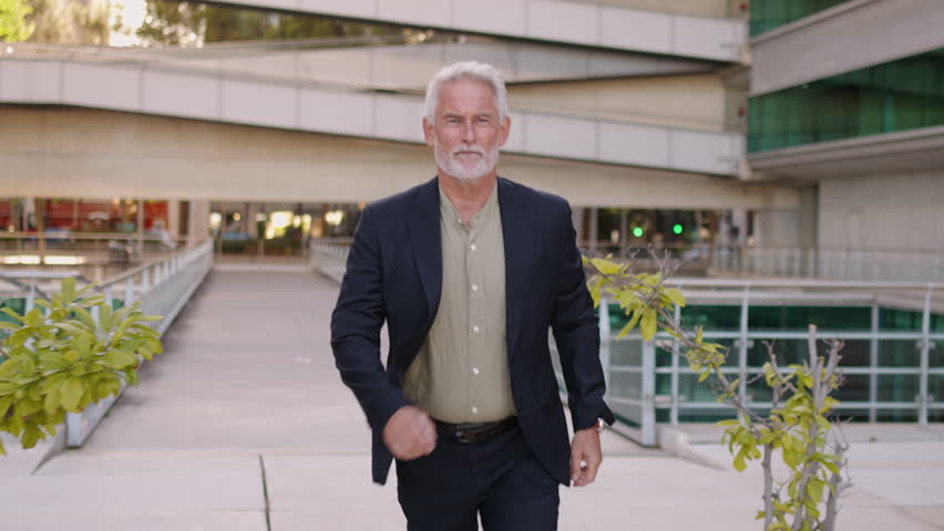 Senior businessman walking while checking time on wristwatch, expressing urgency and professionalism near modern office building