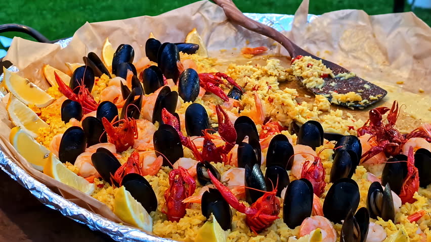A close-up shot of a huge, freshly cooked pan of traditional Spanish seafood paella with saffron rice, mussels, crayfish, and lemon wedges at a food festival.