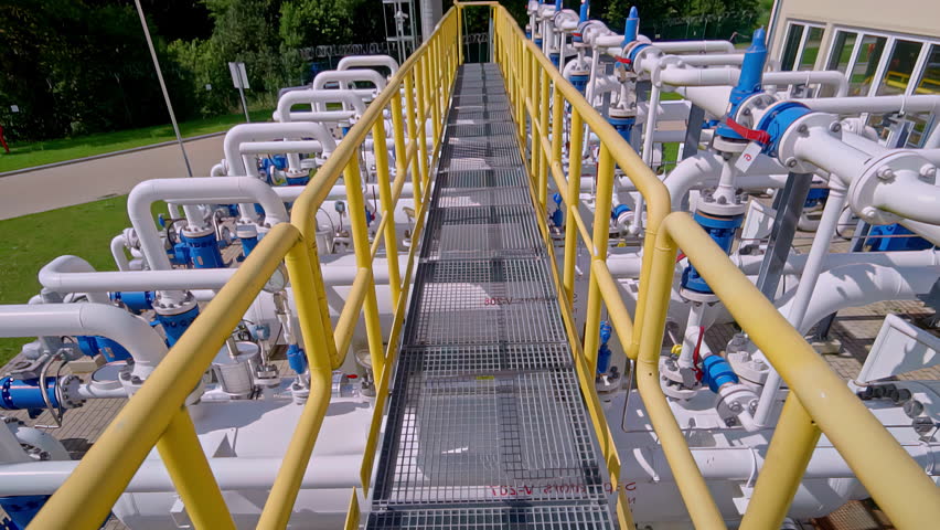 Metal catwalk between rows of pipes and blue valves at a modern natural gas pumping station and transportation facility in Latvia, revealing the infrastructure - A dolly-back reveal shot