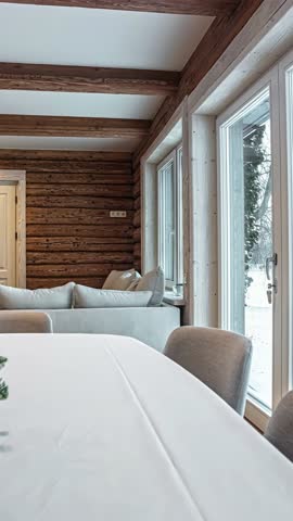 A vertical revealing shot showcases the beautiful interior design of a modern rustic log cabin in Latvia, featuring exposed wooden beams, a cozy fireplace, and a table with Christmas decorations.