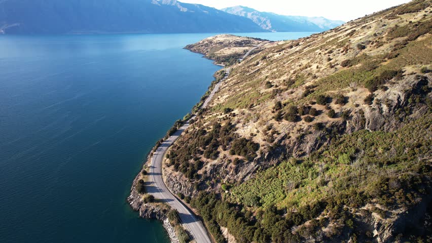 Road Along The Tranquil Blue Waters Of Lake Hawea In Summer In South Island, New Zealand. - aerial shot