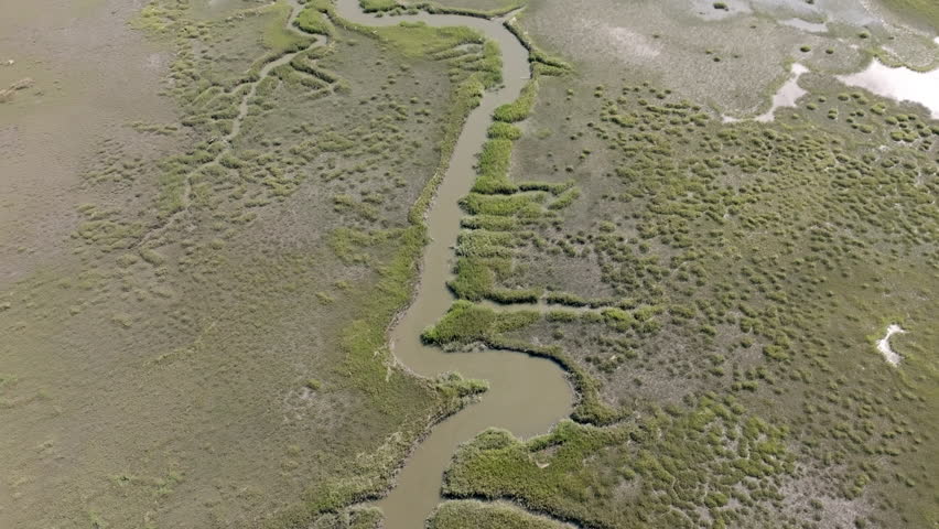 Aerial video of a winding tidal creek cutting through the marshland of Hilton Head Island. The waterway meanders through dense patches of grass and muddy inlets, forming organic patterns.