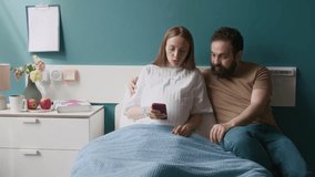 Medium full shot of pregnant young woman sitting in hospital bed with caring husband, viewing baby ultrasound images on smartphone, wife feeling painful contractions, grimacing, gripping man by hand - Powered by Shutterstock - Get 15% off with code: PIKWIZARD15