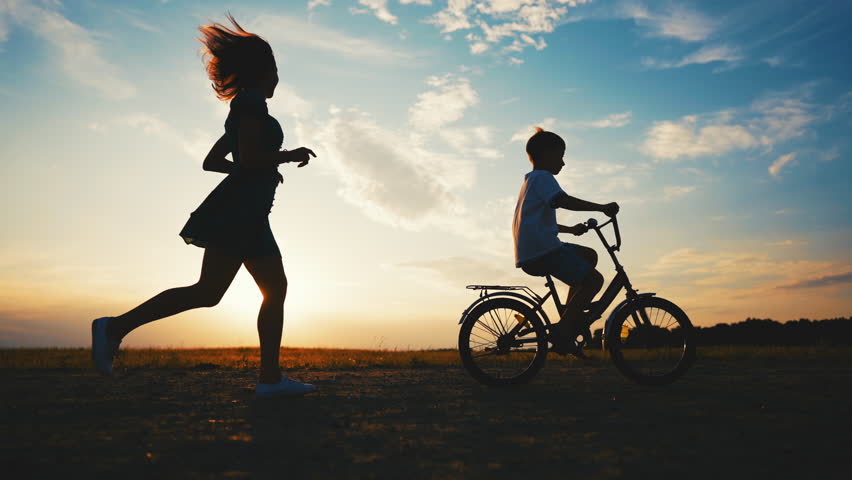 Mother and son in nature, lady running behind her child riding bike, slow motion . Cinematic silhouette shot with mom and little boy in beautiful field, carefree and happy people, childhood moments