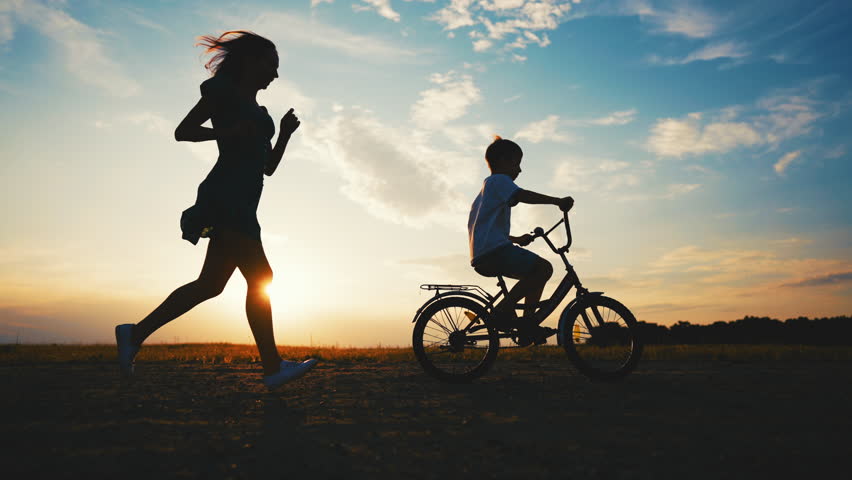 Mother and son in nature, lady running behind her child riding bike, slow motion . Cinematic silhouette shot with mom and little boy in beautiful field, carefree and happy people, childhood moments