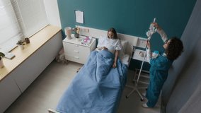 High-angle shot of female nurse setting up bag with intravenous fluid on drip stand for young pregnant woman, lying in bed with smartphone while receiving infusion, then tucking in patients blanket - Powered by Shutterstock - Get 15% off with code: PIKWIZARD15