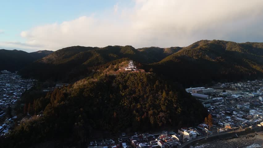 Aerial view of Gujo Hachiman Castle perched atop a hill, overlooking a town nestled amidst rolling mountains under a sky, Gujo Hachiman, Gifu, Japan.