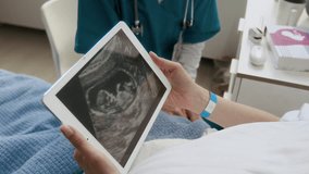 Medium close-up of hands and face of loving young pregnant Caucasian woman lying in bed in maternity ward, admiring ultrasound image of fetus on tablet, while chatting to female midwife - Powered by Shutterstock - Get 15% off with code: PIKWIZARD15
