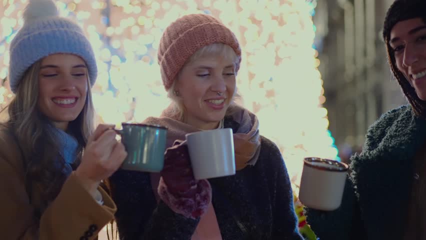 Slow motion close-up of three happy young women clinking mugs of hot mulled wine or chocolate, celebrating together at a festive Christmas fair at night.