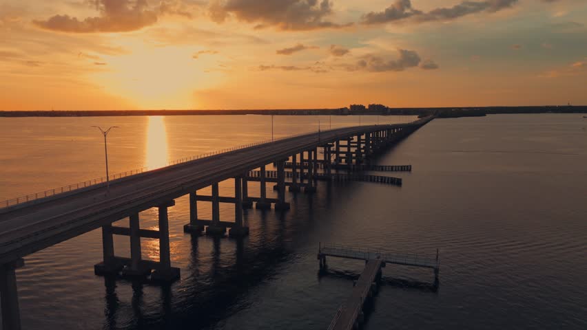 Aerial wide shot of Caloosahatchee bridge leading to North Fort Myers city in background. Golden sunset time in Florida. Orange colored sky. Silhouette of construction.