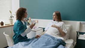 Medium full shot of Caucasian female nurse in blue robe placing electronic thermometer against forehead of young pregnant patient to check body temperature during morning rounds at maternity hospital - Powered by Shutterstock - Get 15% off with code: PIKWIZARD15