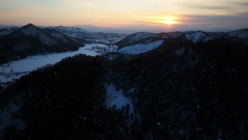 Aerial view of snow-covered mountains and a frozen lake under a pastel sky, creating a serene winter landscape, Niijukuchiku, Yamagata, Japan.