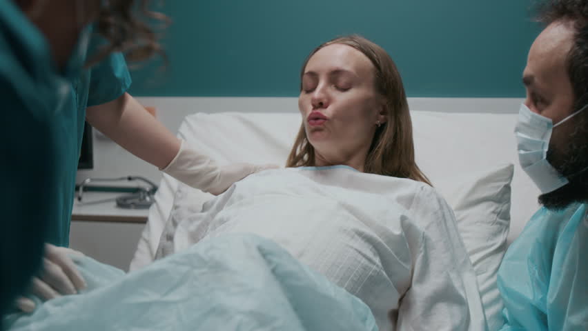 Medium shot of exhausted Caucasian pregnant woman screaming in intense pain during labor, pushing, screaming, then midwife pulling out newborn baby, husband in gown and mask supporting wife