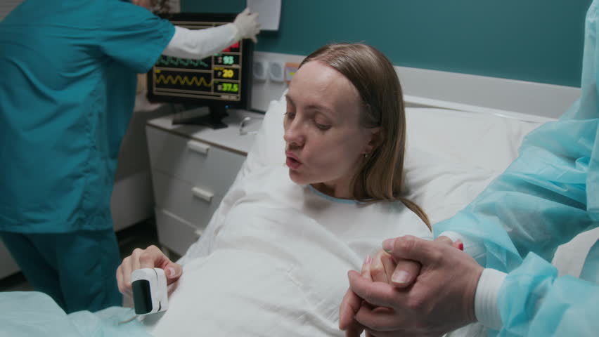 Zoom-out shot of pregnant woman lying on bed in delivery room at maternity clinic, experiencing painful labor contractions, breathing, listening to instructions from midwife, preparing for baby birth