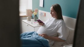 Medium full shot of pregnant young woman in white robe reclining on pillows in bed at maternity ward, recording voice message on smartphone, while sharing news with husband, friend or family - Powered by Shutterstock - Get 15% off with code: PIKWIZARD15