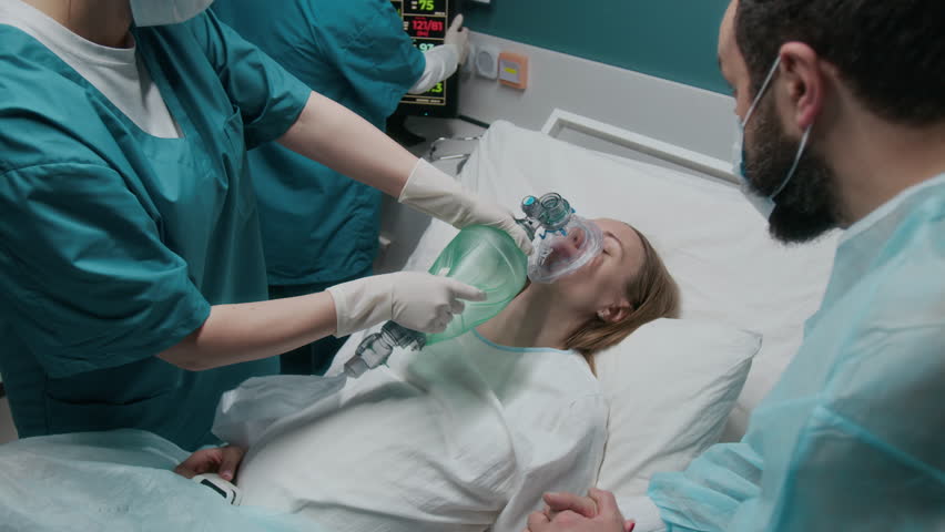 Medium arc shot of maternity hospital ventilating pregnant woman, fainted from pain during labor contractions, using handheld Ambu bag, midwife checking monitors and stressed husband holding her hand