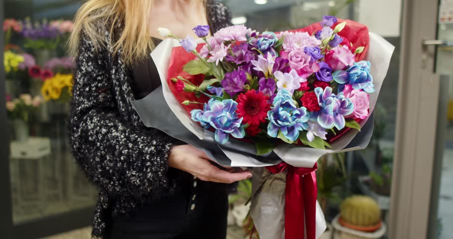 A young woman florist holds a large, colorful bouquet featuring prominent pink roses and blue hydrangeas, wrapped in dark paper. Her hands gently support the arrangement; her face is not visible.