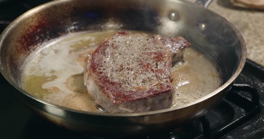 A closeup of thick cut of meat is sizzling in a stainless steel pan on a stovetop, surrounded by rendered fat