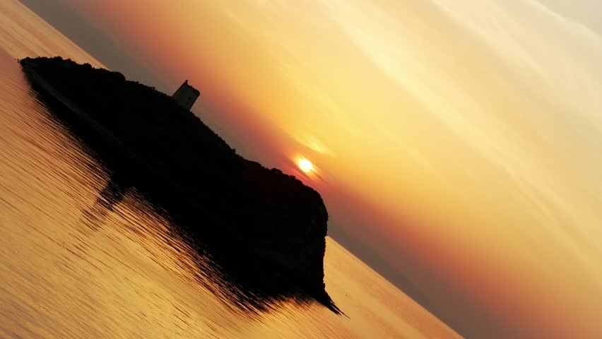 Silhouetted watchtower on islet near Pula at golden sunset, sun glows over calm Mediterranean Sea, romantic and beautiful scenery in Sardinia, Italy. Aerial drone, copy space