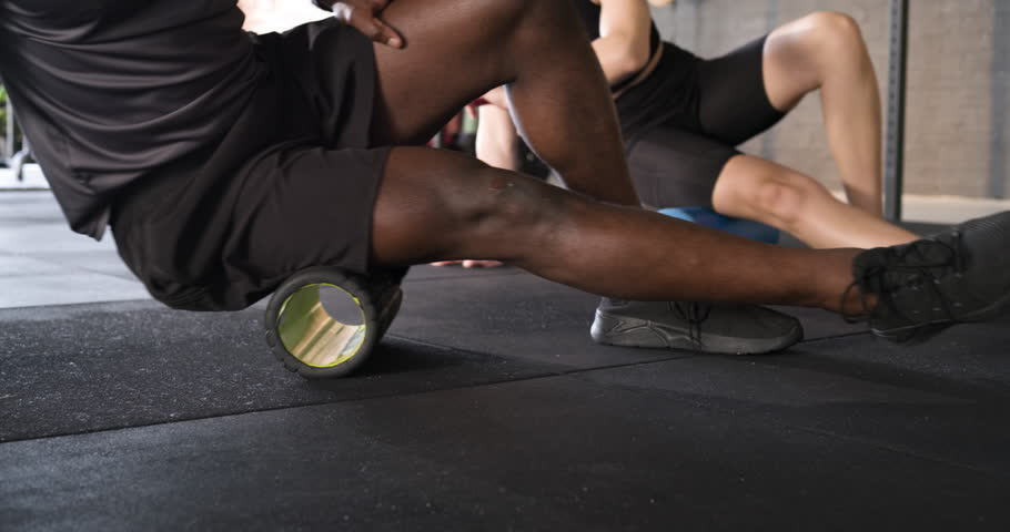 Placing foam rollers under thighs, diverse friends rolling for muscle release at gym, copy space. Fitness, relaxation, recovery, exercise, athletic, wellness, stretching