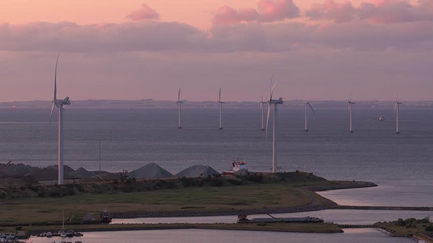 A serene aerial view of Copenhagen during sunset, featuring a wind turbine in motion, industrial buildings, storage tanks, and the calm waterfront.