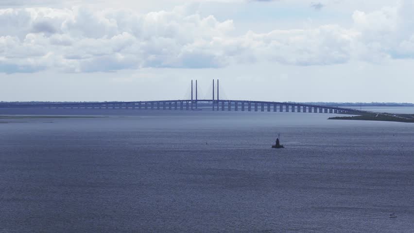 A serene aerial view of the Oresund Bridge connecting Denmark and Sweden, with a small vessel moving across calm blue waters under a partly cloudy sky.