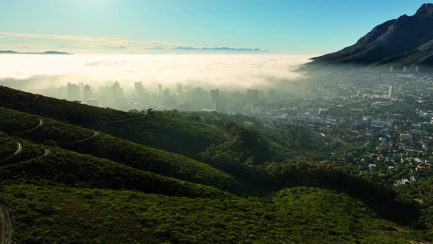 Aerial view of the city skyline shrouded in ethereal fog, juxtaposed against the lush green mountains, a captivating blend of nature and urban landscape, Cape Town, Western Cape, South Africa.