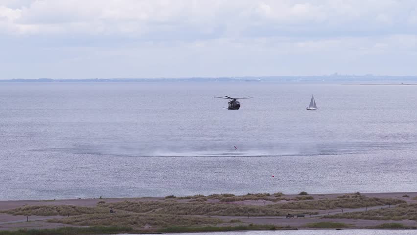 A military helicopter flies low over water, creating ripples. Sailboats, a sandy shoreline, and a walking path with people are visible under a cloudy sky.