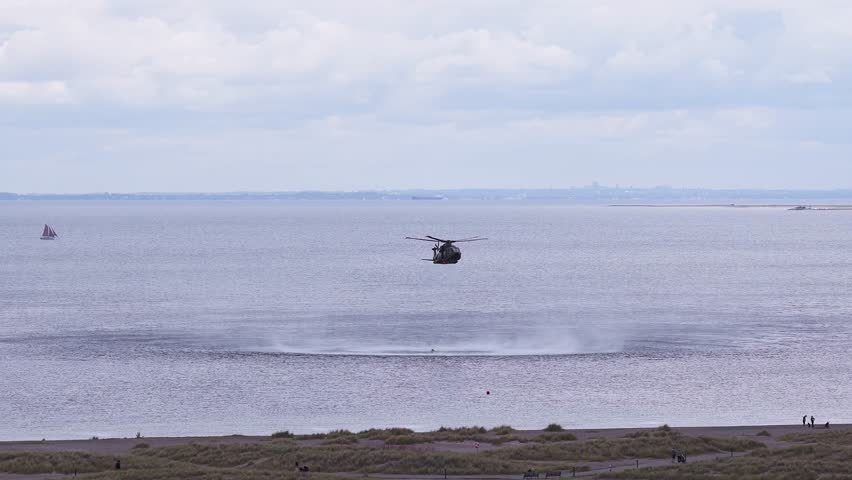 A military helicopter hovers low over water, creating ripples. A sailboat and city skyline are visible, with pedestrians on a grassy shoreline.