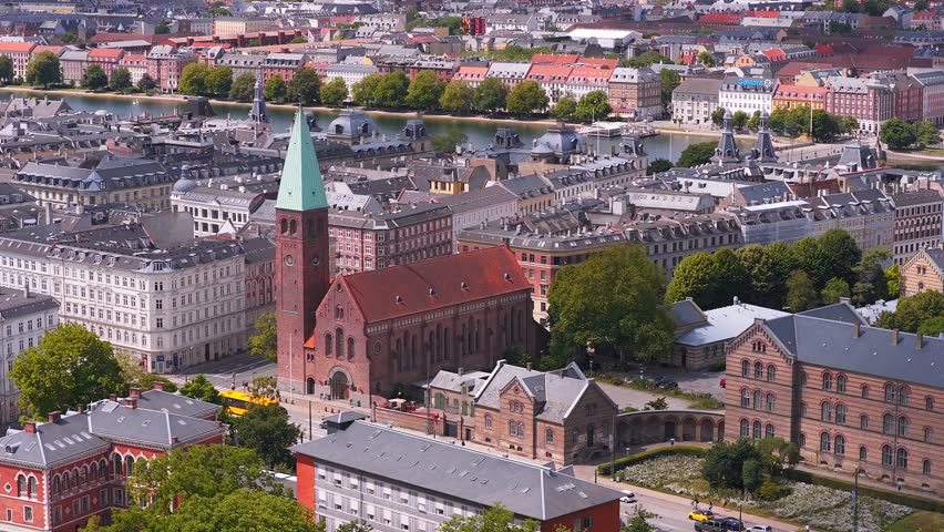 Aerial view of Copenhagen, Denmark, with historic buildings, green spaces, and a body of water in the background. Slow pan across the cityscape.