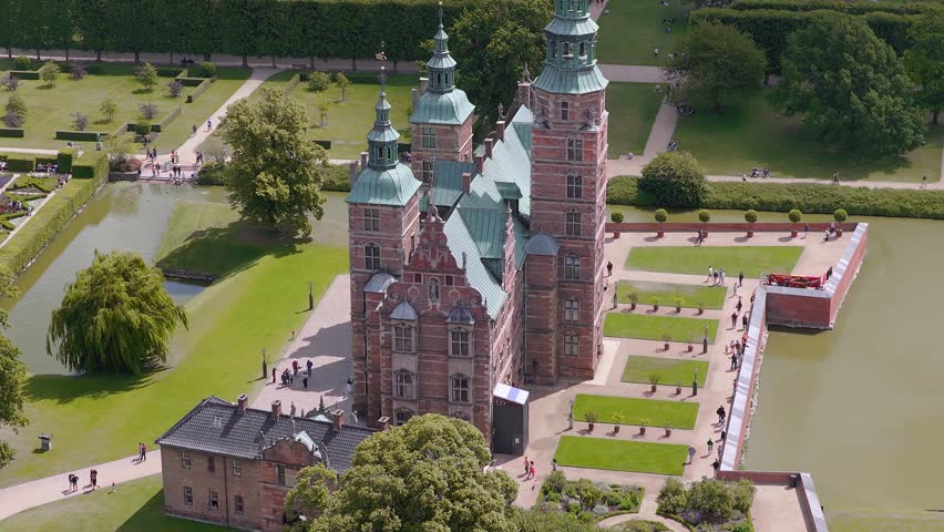 Aerial view of Rosenborg Castle in Copenhagen, Denmark, with red brick facade, green copper roofs, lush gardens, a moat, and light pedestrian activity.